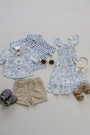 Flat lay of two boys button-up shirts in light blue patterns, beige shorts, and a girls dress with ruffled sleeves, paired with sunglasses and sandals, showcasing a stylish summer outfit.