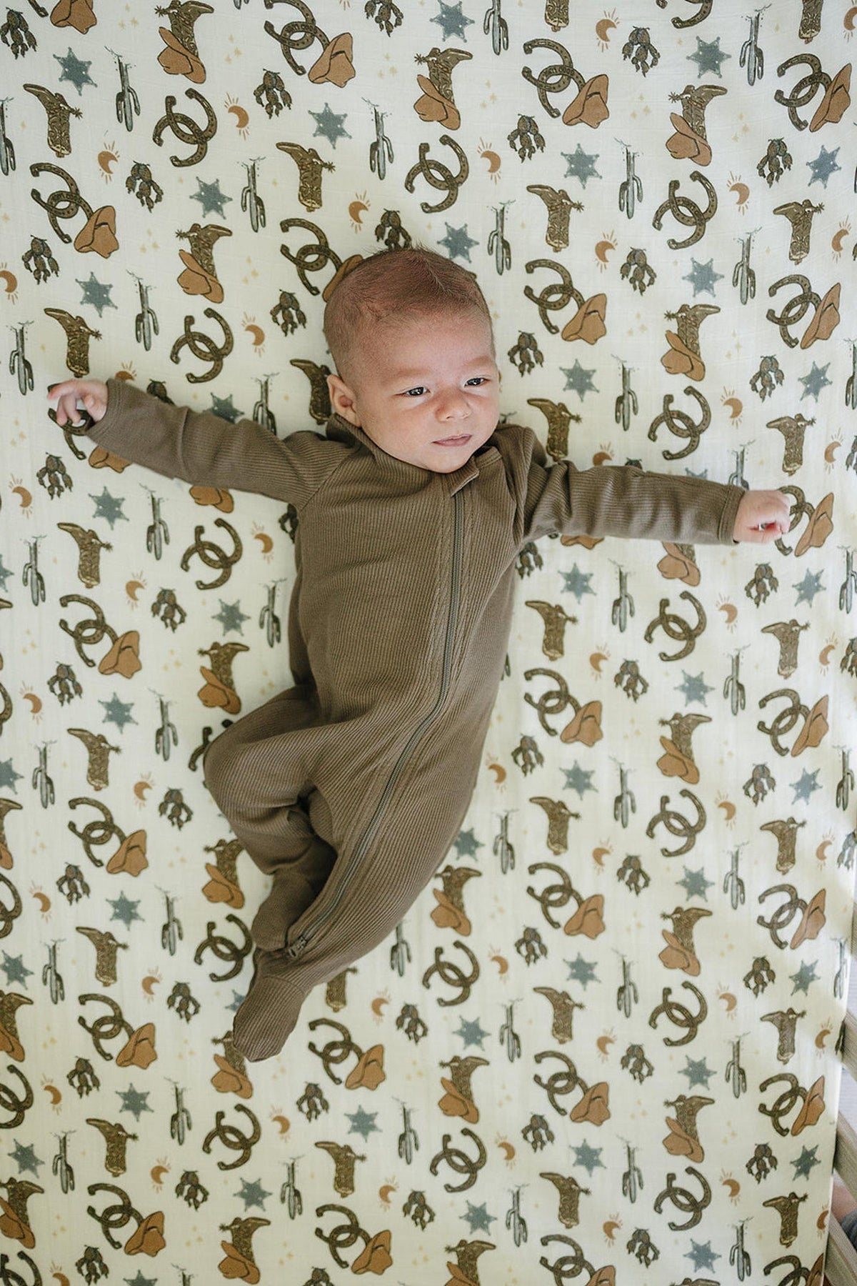 A baby in a brown onesie lies on a cowboy-themed muslin crib sheet with hats and stars pattern.
