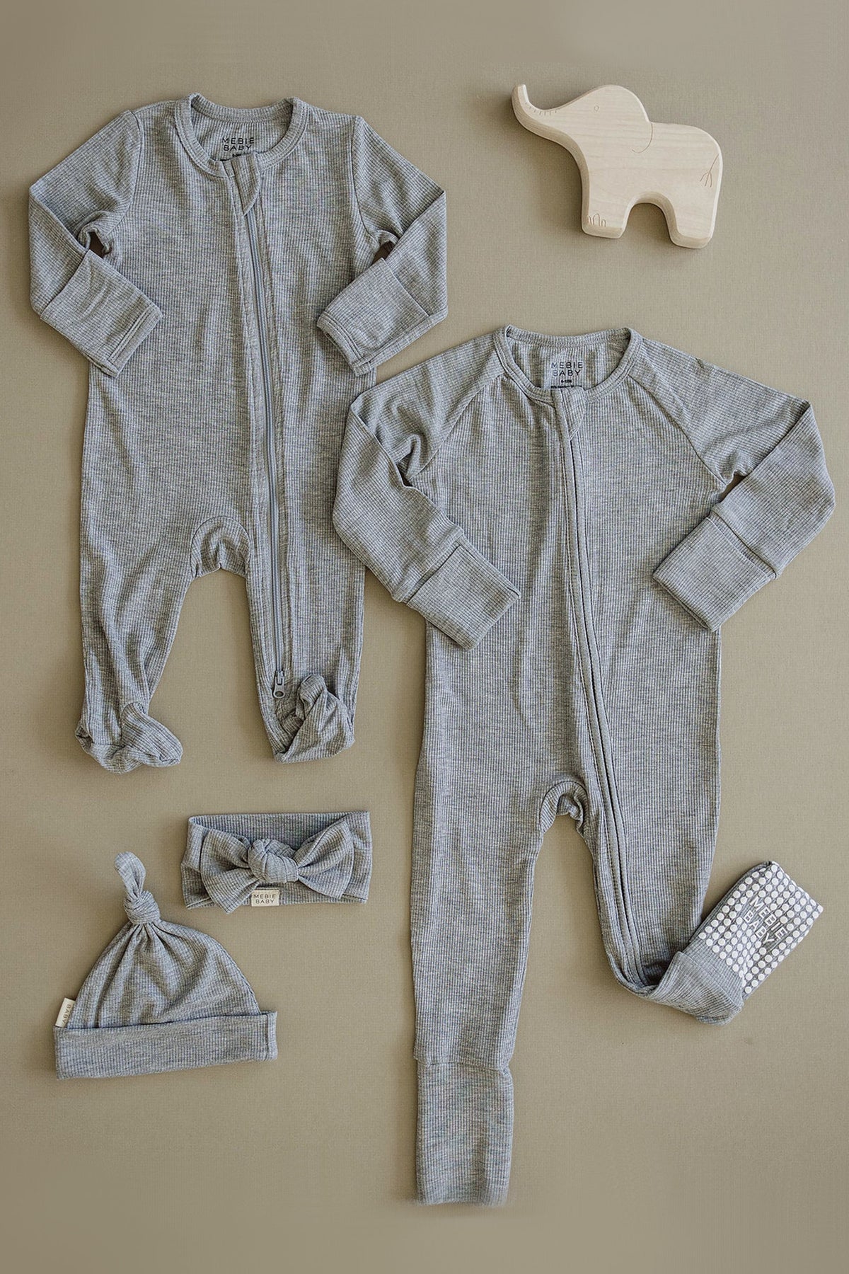 Two heather grey ribbed zipper pajamas with matching hats and headband, displayed on a beige surface alongside a wooden elephant.