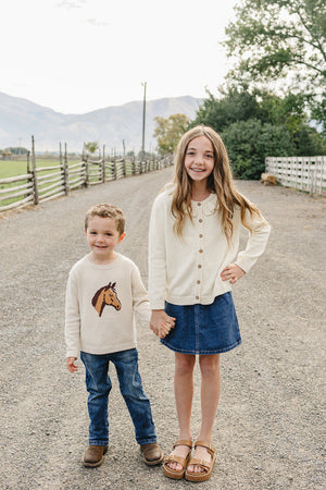 A young boy and girl stand together on a gravel path, with the boy wearing a cream sweater featuring a horse design and the girl in a button-up cream cardigan.