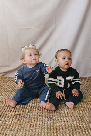 Two infants sit on a woven rug, one wearing a dark green cotton set with white stripes, and the other in a blue outfit. Both have a relaxed, playful expression.