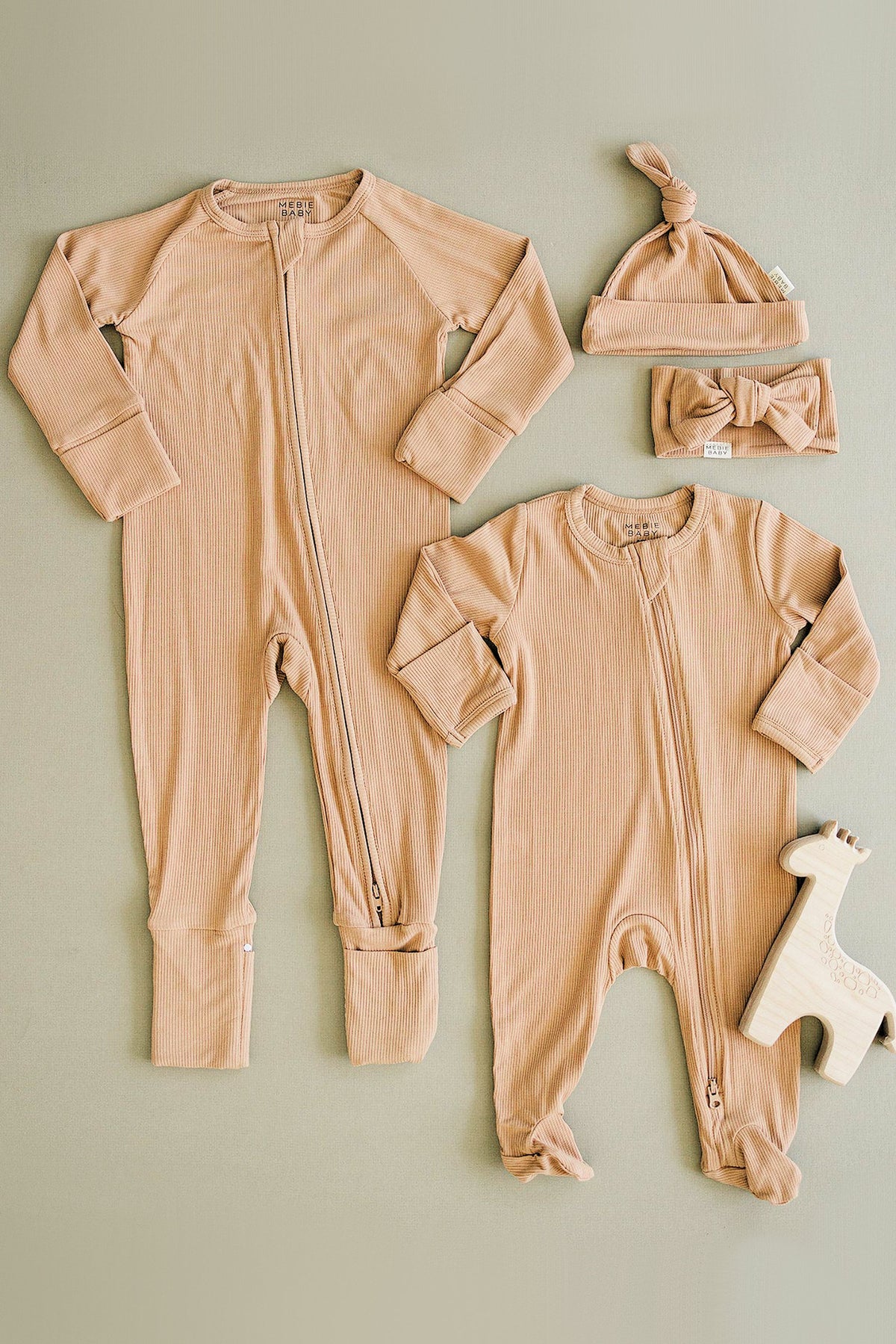 Two latte-colored ribbed bamboo baby pajamas with zippers, matching hat, headband, and a wooden giraffe toy.