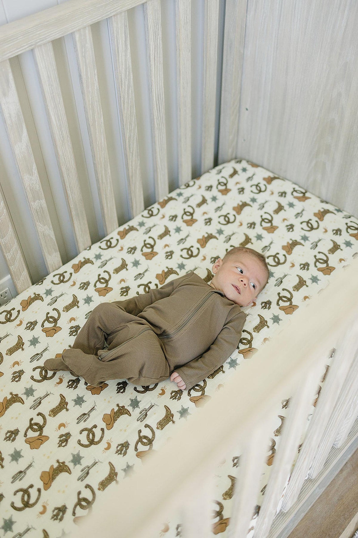 A baby in a brown outfit lies on a crib sheet with cowboy-themed prints in a wooden crib.