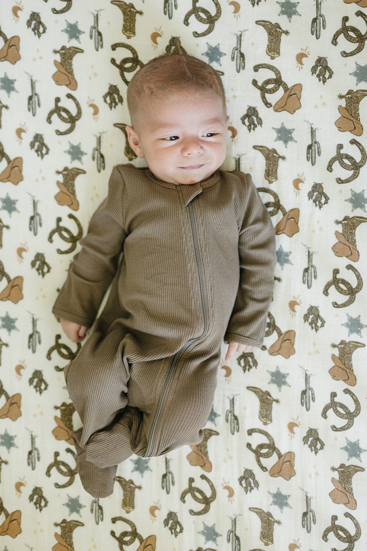 A baby in a brown outfit lies on a cowboy-themed muslin crib sheet with horseshoes and stars.