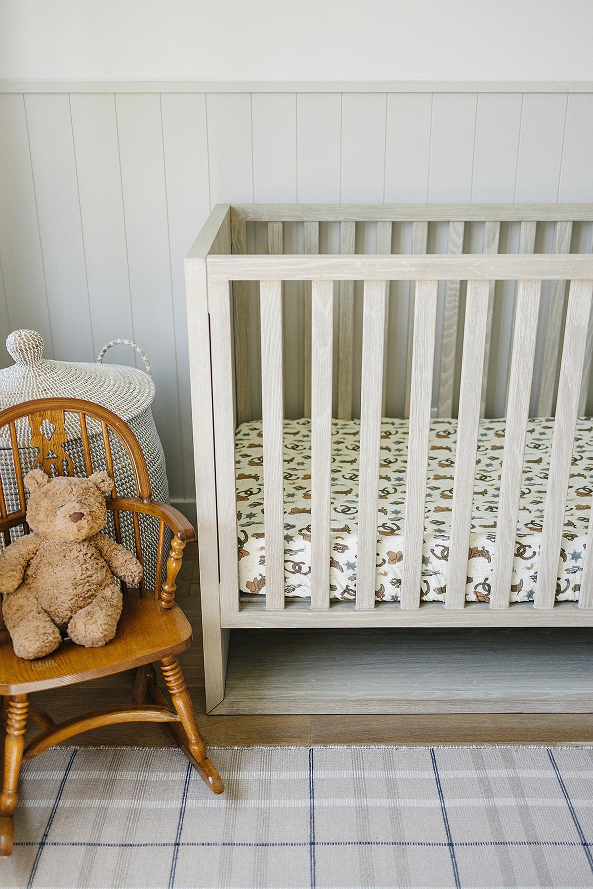A wooden crib with a cowboy-themed muslin sheet, next to a teddy bear on a wooden chair.