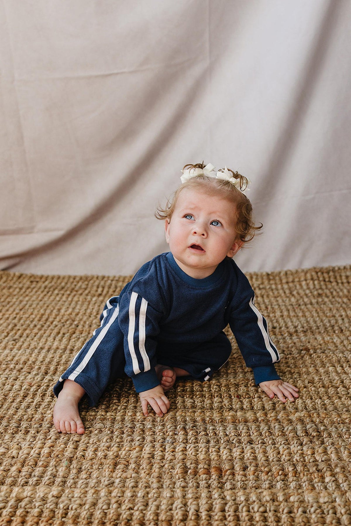 A toddler wearing a navy terry cloth tracksuit with white stripes crawls on a textured, natural fiber rug. The child has curly hair adorned with a small bow, gazing upward.