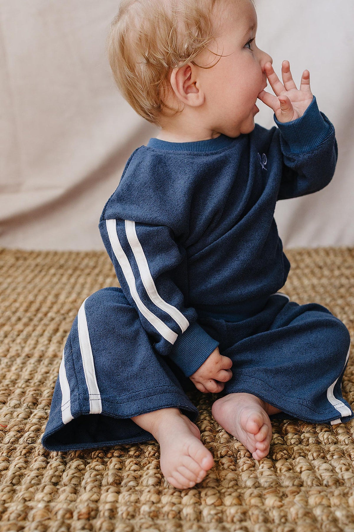 A toddler in a navy terry cloth tracksuit with white stripes sits on a textured, natural fiber rug, playfully chewing on their hand.