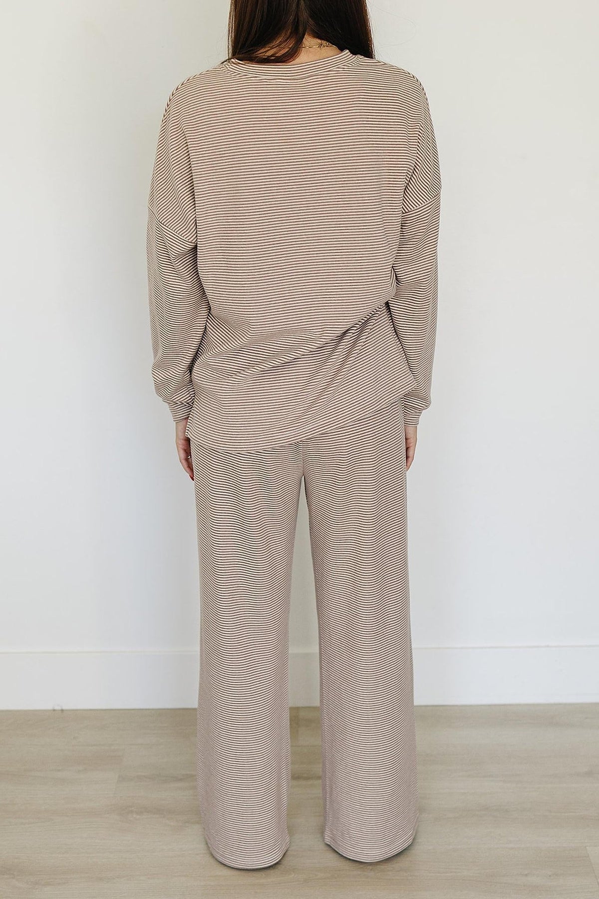 A woman is seen from behind, wearing a beige striped loungewear set made of soft cotton. The outfit features a relaxed fit, perfect for comfortable lounging at home.
