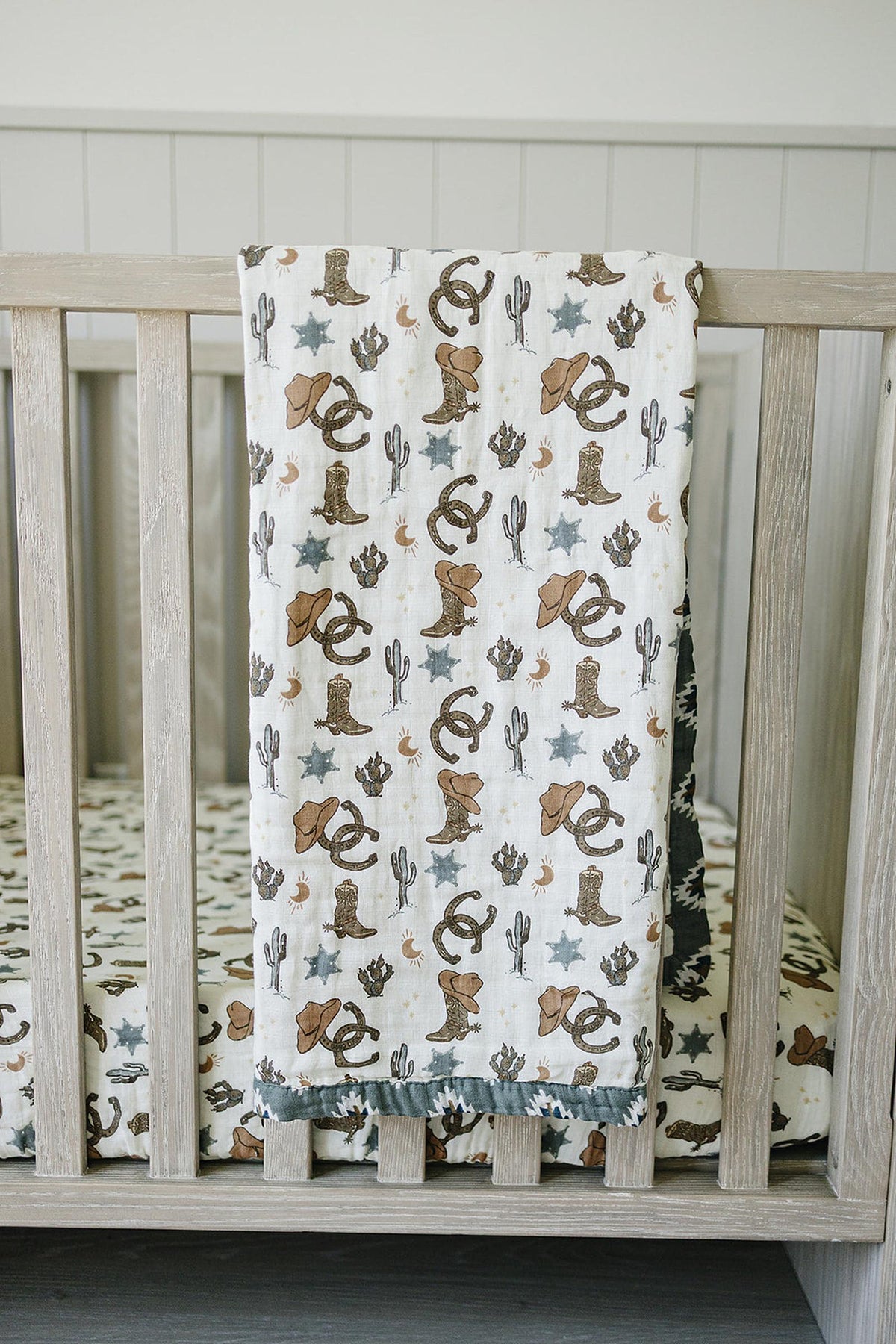 A muslin crib sheet with cowboy-themed prints, including hats, boots, and cacti, draped over a wooden crib.