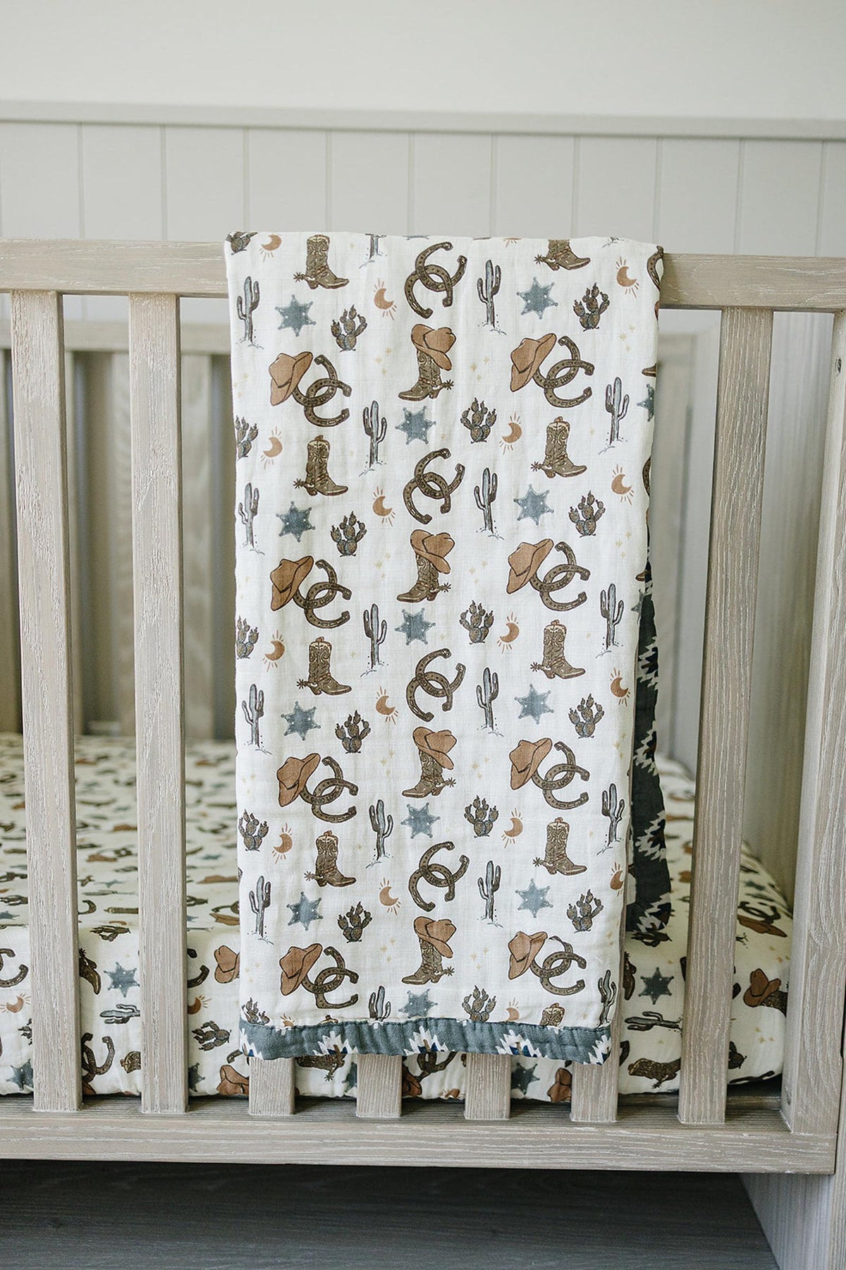 A muslin quilt with cowboy-themed prints, including boots and hats, draped over a wooden crib.