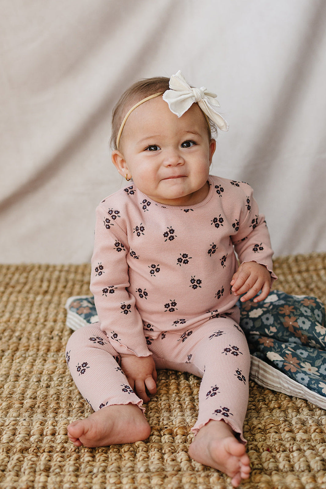 A baby sits on a woven rug, wearing a cozy pink cotton outfit adorned with small floral patterns. A white bow headband complements the look, creating a sweet, playful style.