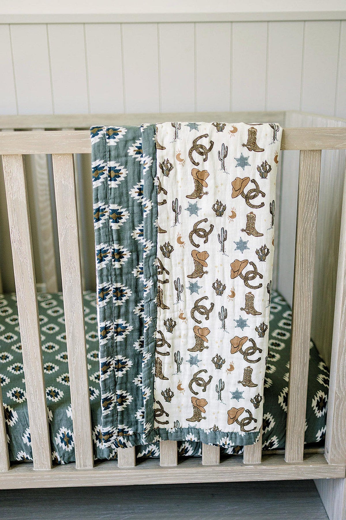 Two muslin quilts hang on a crib, featuring Aztec and cowboy-themed patterns in neutral colors.