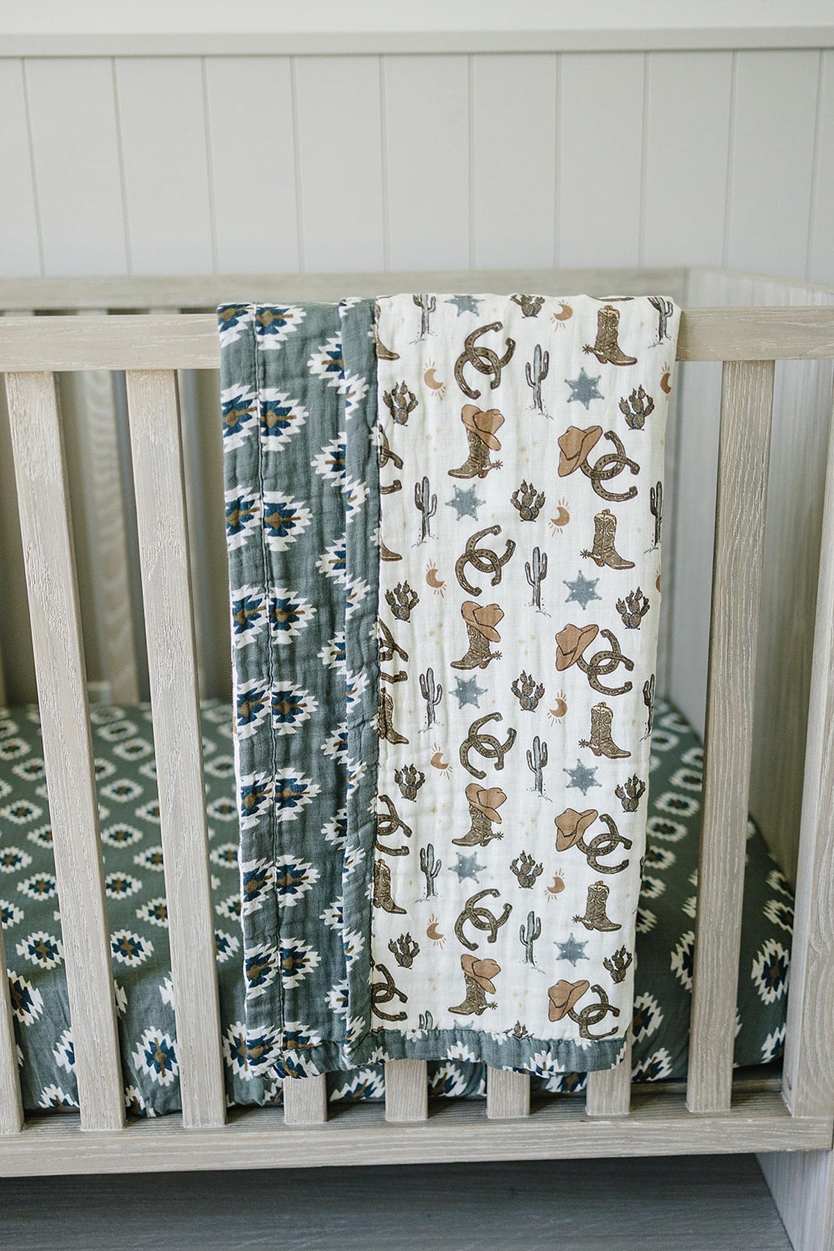 Crib with two muslin blankets draped over the side, featuring Southwest Aztec patterns and cowboy-themed prints.