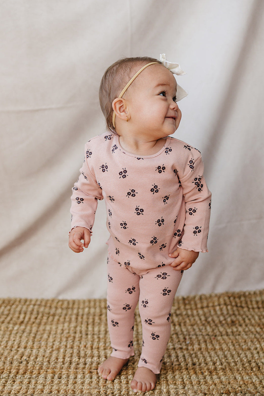 A smiling baby wearing a pink cotton outfit featuring small floral patterns stands on a woven rug. The outfit is comfortable and perfect for play or relaxation.