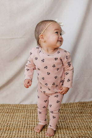 A smiling baby wearing a pink cotton outfit featuring small floral patterns stands on a woven rug. The outfit is comfortable and perfect for play or relaxation.