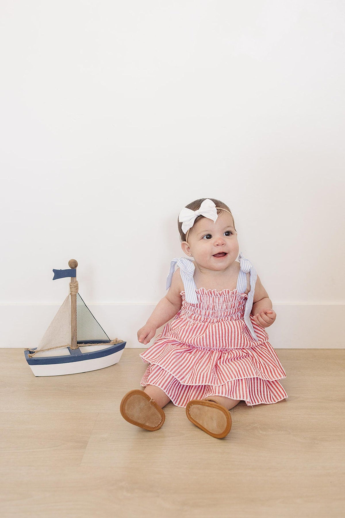 Baby wearing a red striped ruffle dress and white bow headband, sitting beside a small wooden sailboat.