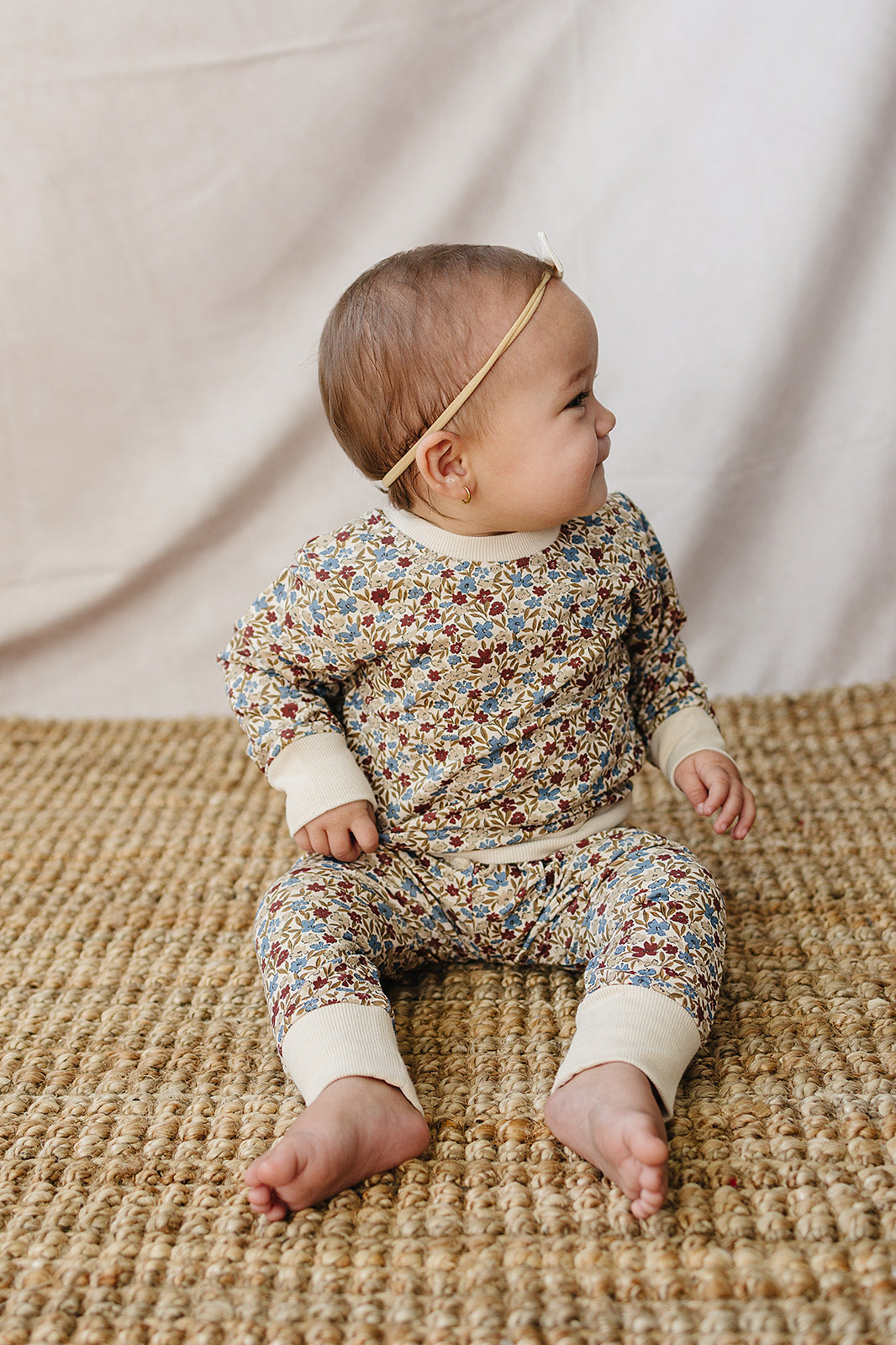 A baby sits on a woven rug, wearing a soft, floral-printed two-piece set in muted colors. The outfit features a relaxed fit and is made from breathable cotton for comfort.