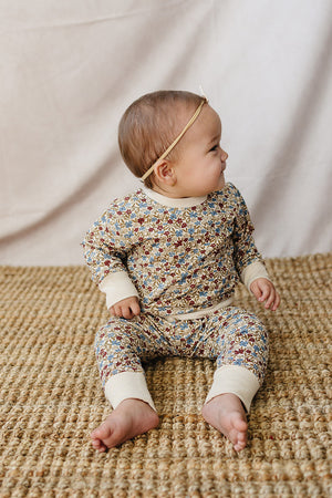 A baby sits on a woven rug, wearing a soft, floral-printed two-piece set in muted colors. The outfit features a relaxed fit and is made from breathable cotton for comfort.