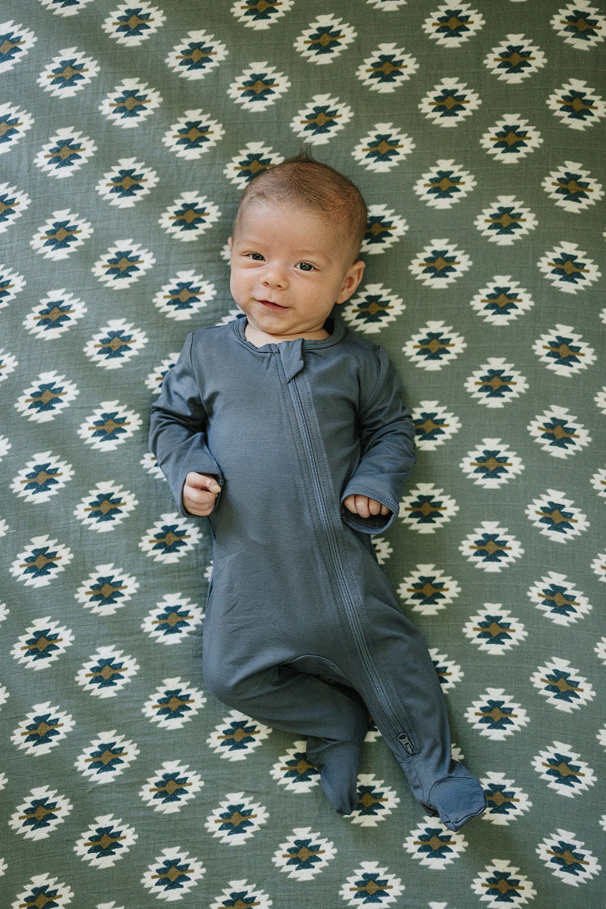A baby in a blue onesie lies on a green Southwest Aztec patterned crib sheet, smiling contentedly.