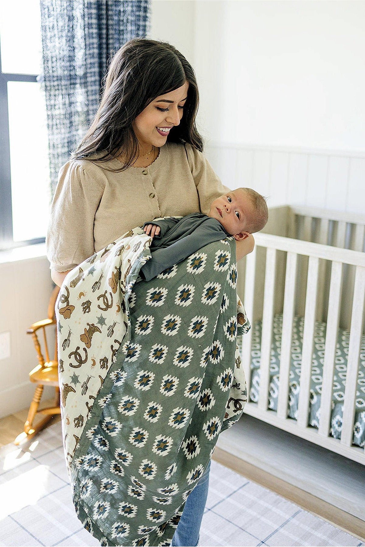A woman holds a baby wrapped in a green and white Aztec-patterned muslin quilt, standing by a crib.