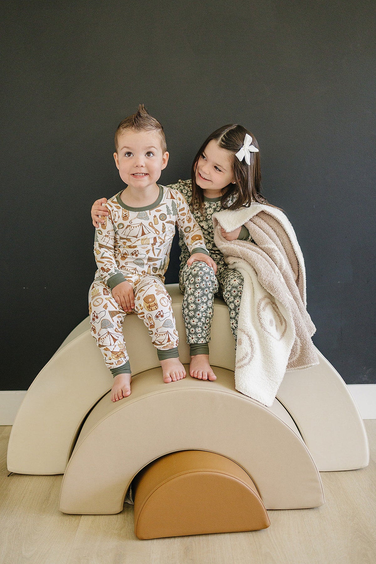 Two children wearing camping-themed pajamas sit on beige play blocks, one holding a cozy blanket.