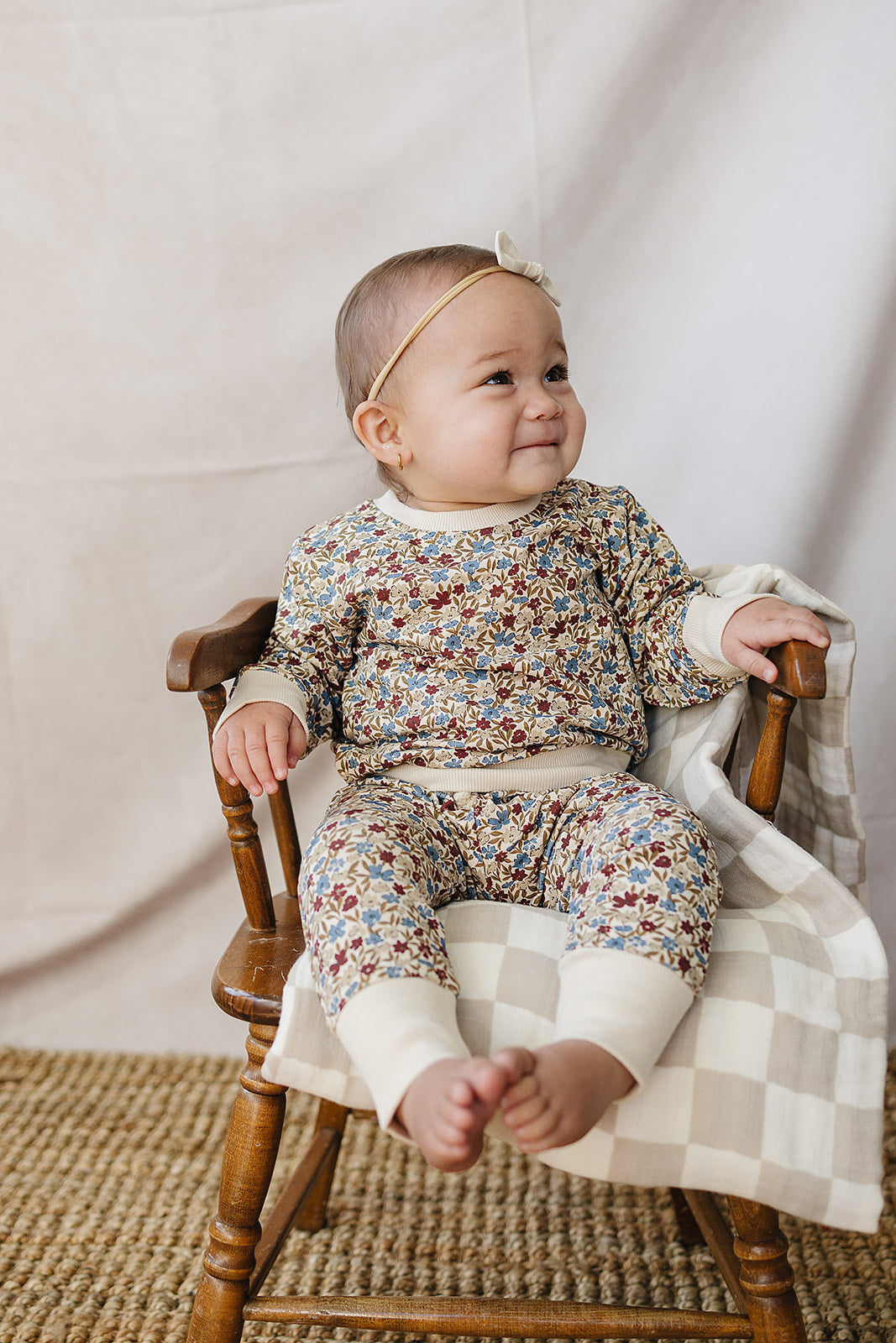 A smiling baby sits in a wooden chair, wearing a soft, floral-patterned two-piece set made from breathable cotton. The outfit features a relaxed fit, perfect for comfort and play.
