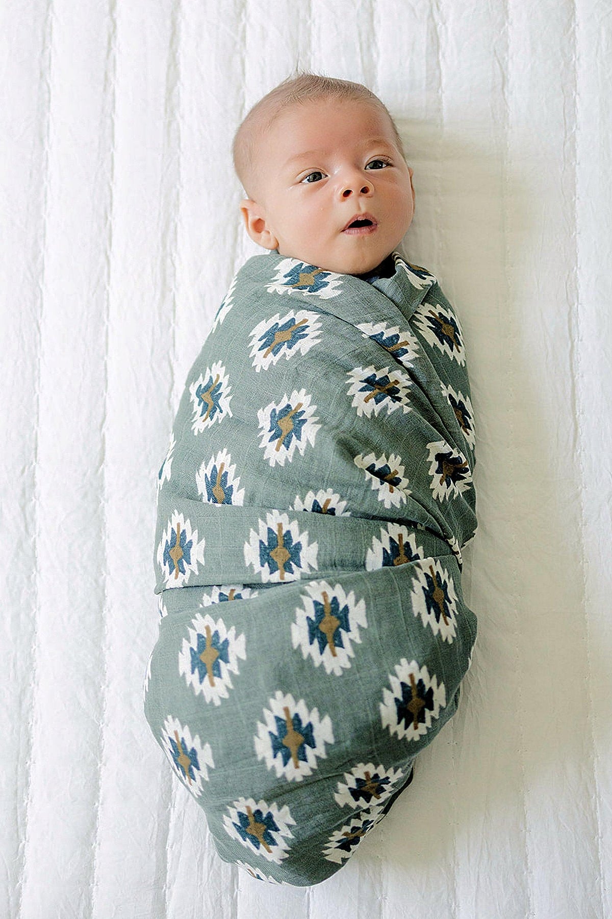 A baby is swaddled in a green muslin blanket with a Southwest Aztec pattern, lying on a white surface.