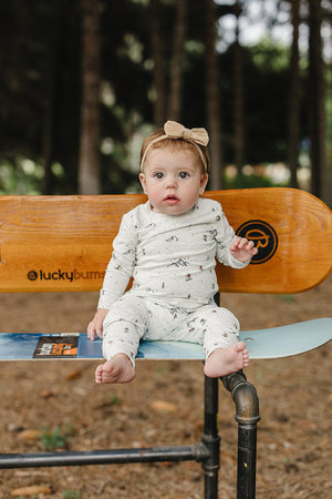 A baby sits on a wooden bench, wearing a cozy, light-colored pajama set with a ski print and a bow.