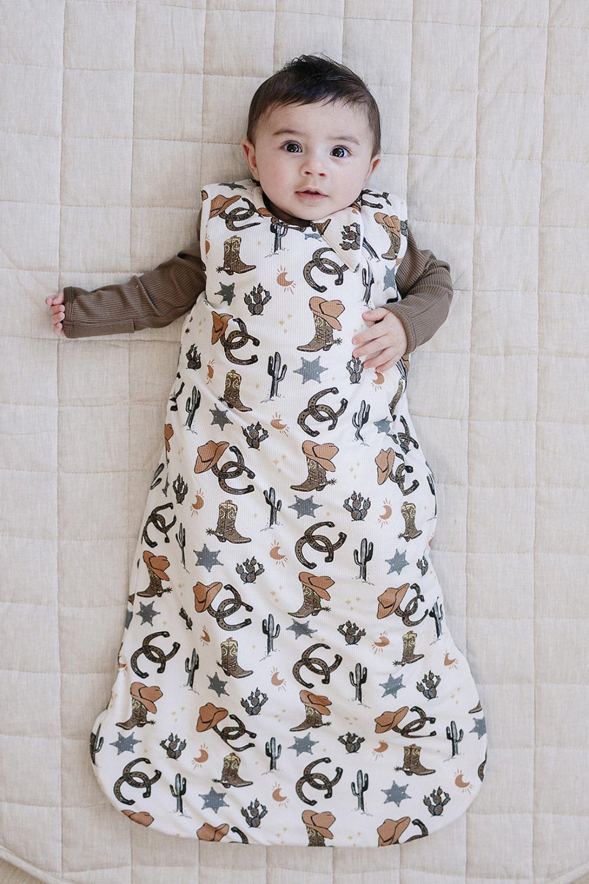 A baby lies on a light quilted surface, wearing a bamboo sleep sack featuring a whimsical cowboy print with cacti and animals.