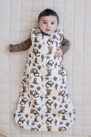 A baby lies on a light quilted surface, wearing a bamboo sleep sack featuring a whimsical cowboy print with cacti and animals.
