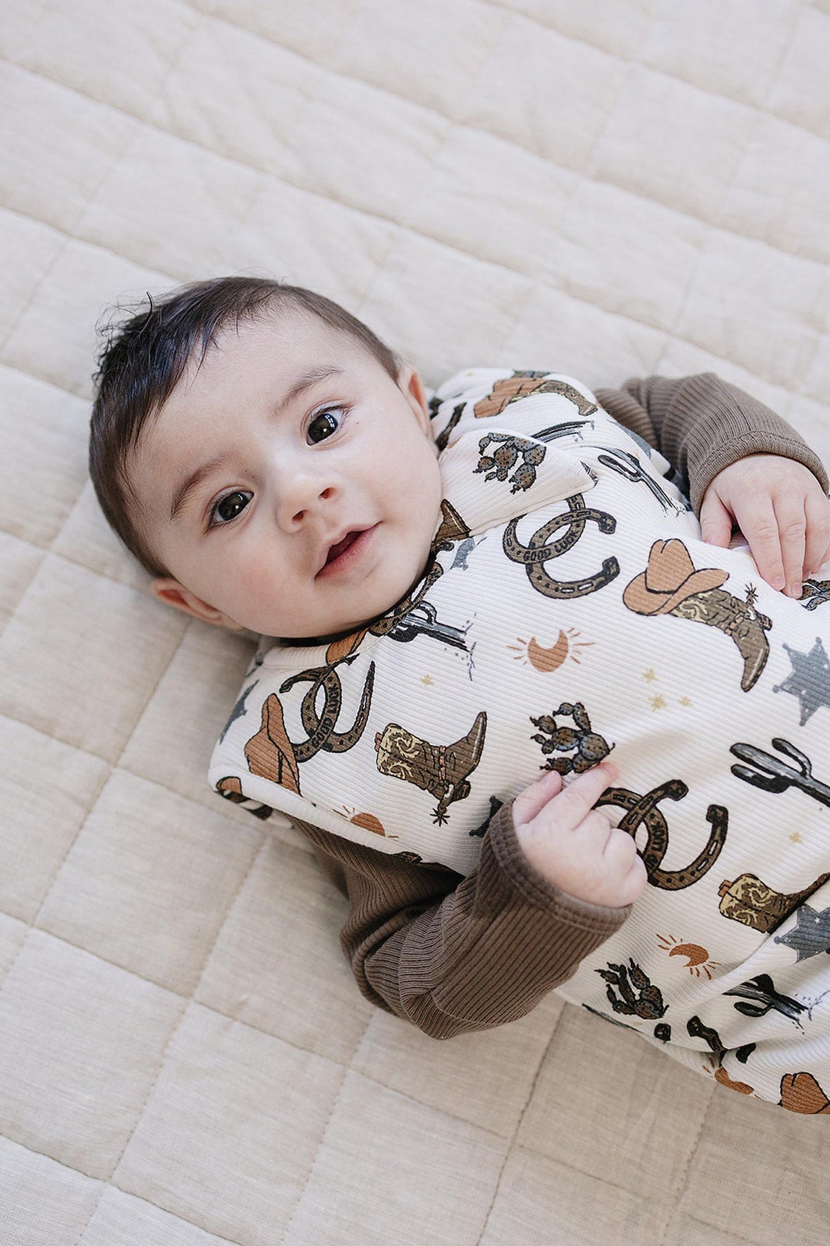 A baby lies on a light-colored quilt, wearing a soft bamboo sleep sack featuring a playful cowboy print with stars, hearts, and western motifs.