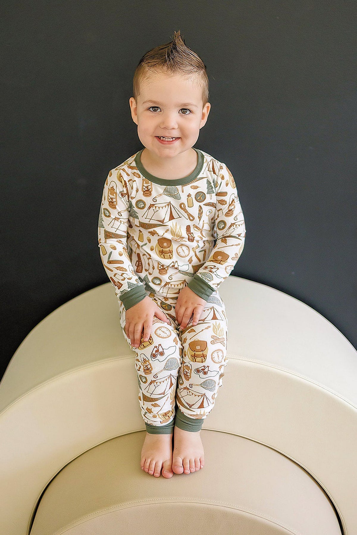 A child wearing a camping-themed pajama set with green trim sits on a beige chair, smiling.