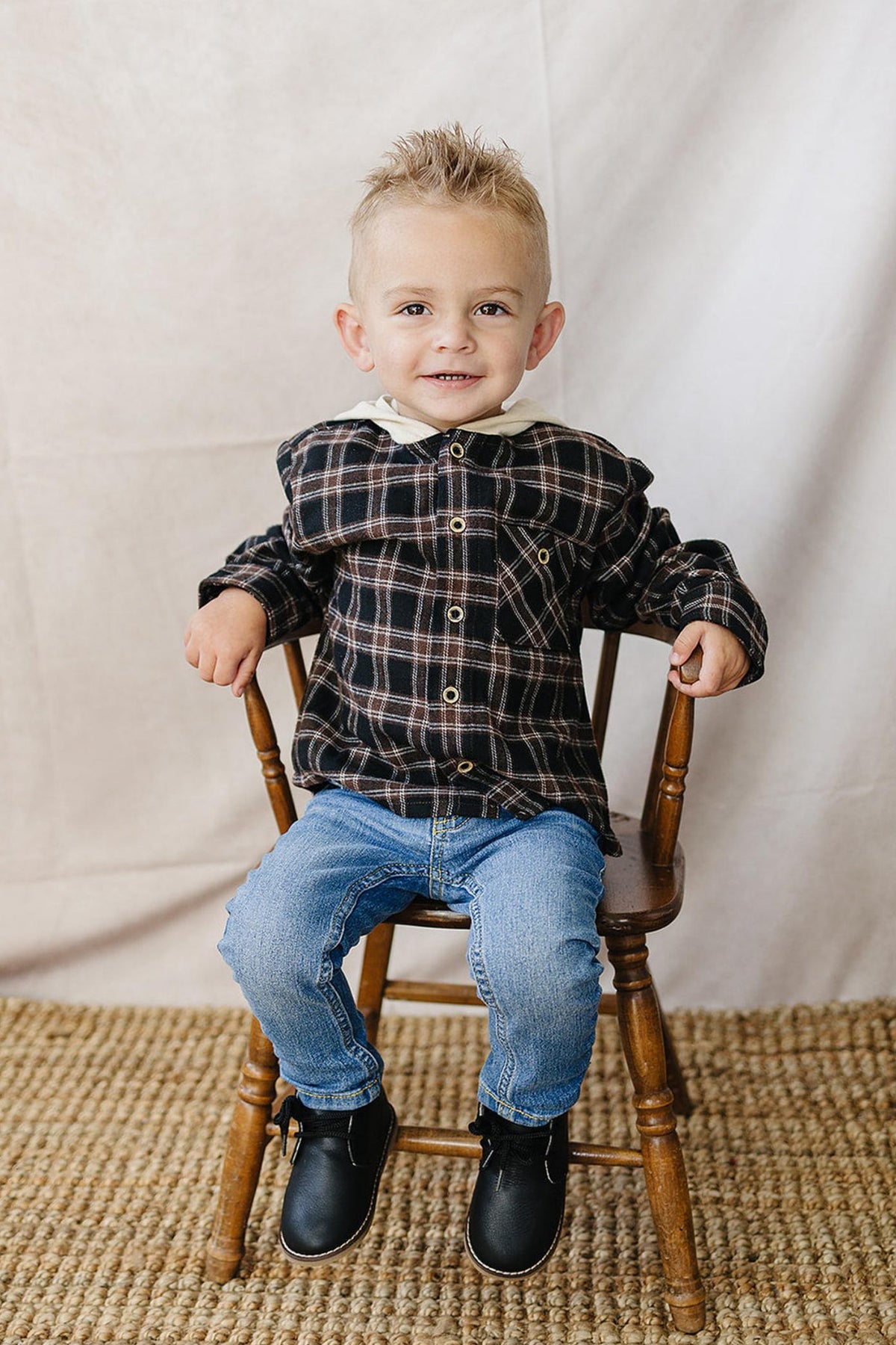A smiling young boy sits on a wooden chair, wearing a dark plaid hooded button-up shirt, paired with blue jeans and black shoes, against a neutral backdrop.