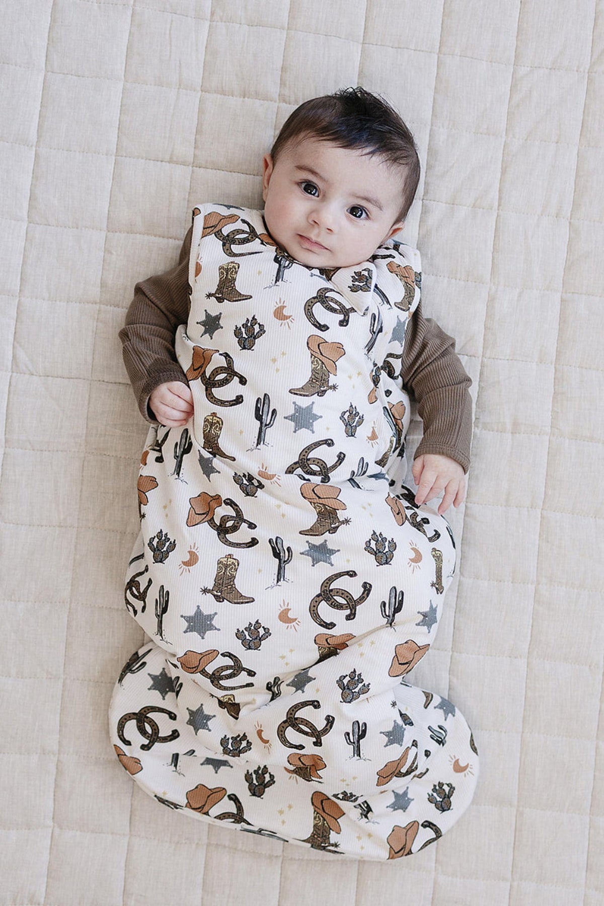 A baby lies comfortably in a bamboo sleep sack featuring a charming cowboy print with hats, cacti, and stars. The soft fabric provides a cozy, breathable layer for little ones.