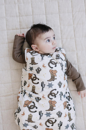 A baby lies on a light beige quilt, wearing a cowboy-themed bamboo sleep sack adorned with boots, stars, and cacti. The infant gazes up, showcasing the soft, breathable fabric.