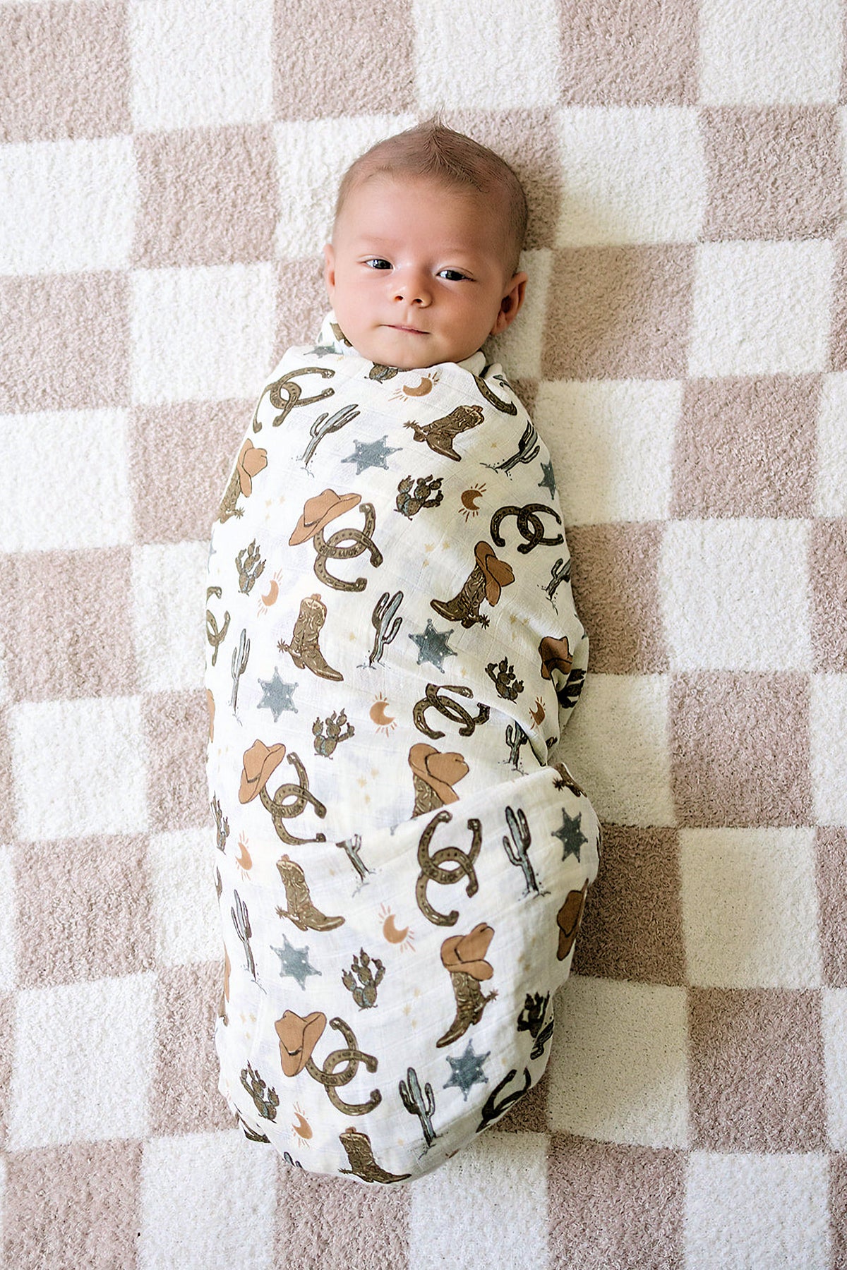 Baby swaddled in a cowboy-themed muslin blanket lies on a checkered beige and white rug.