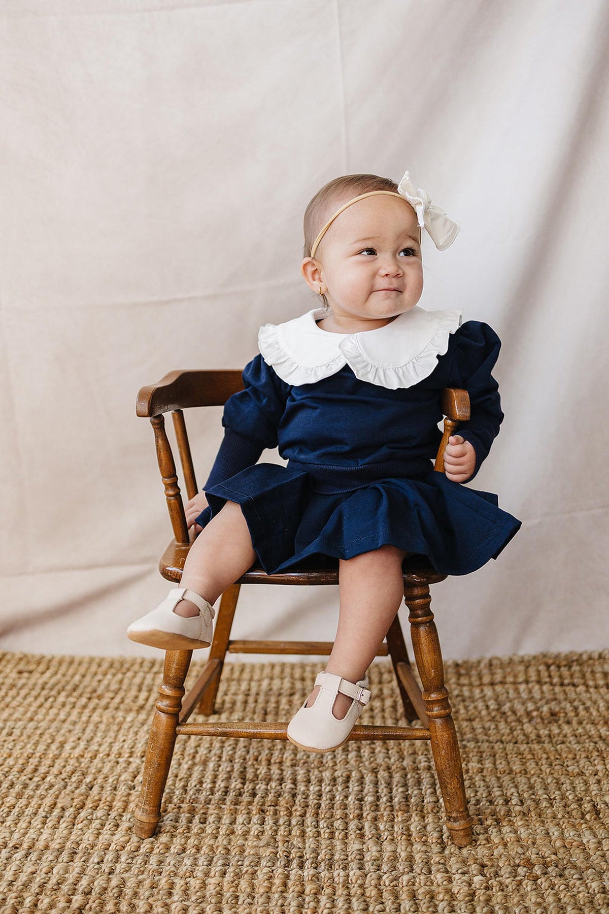 A toddler sits on a wooden chair, wearing a navy dress with a white Peter Pan collar.