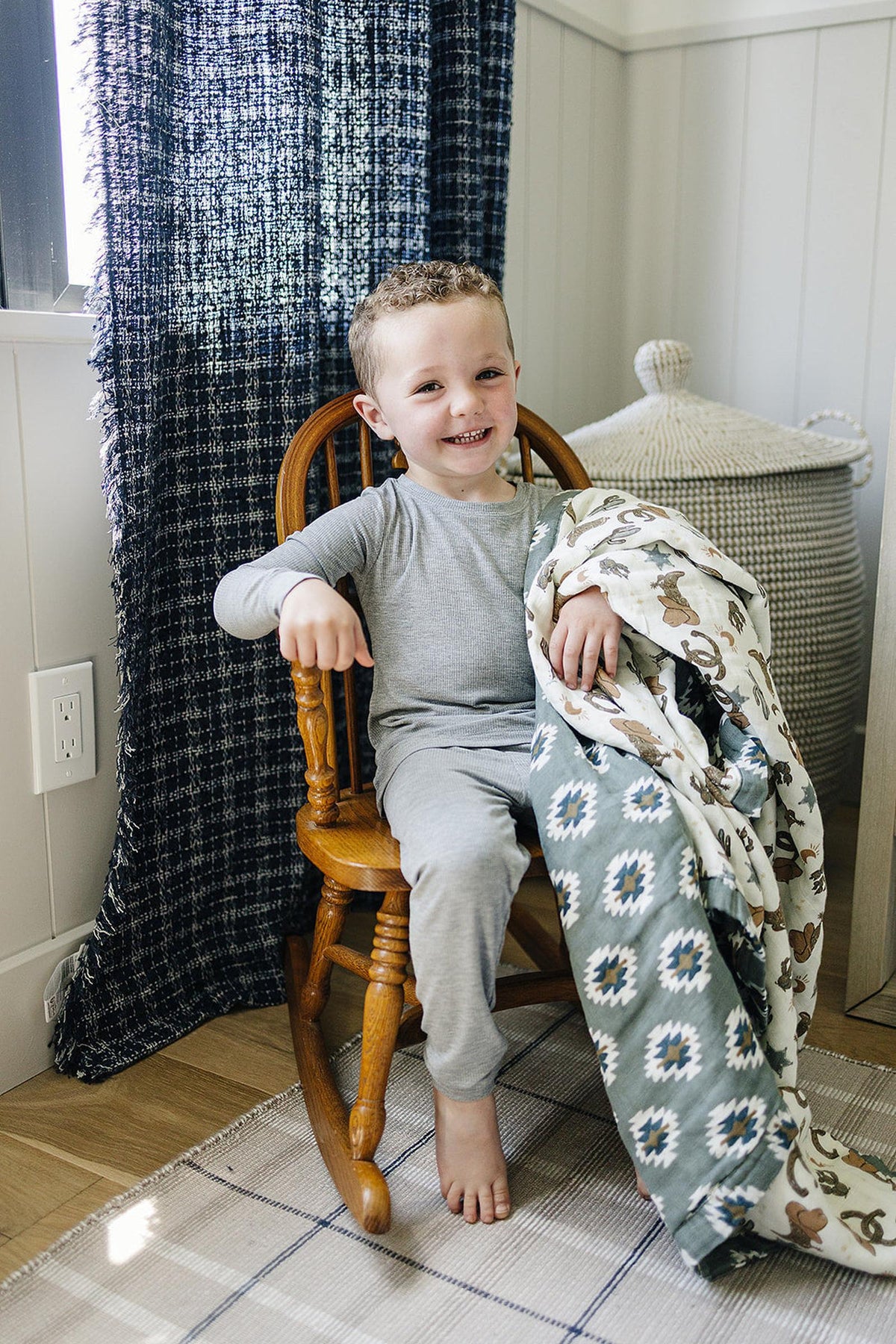 A smiling child in grey pajamas sits on a wooden chair holding a Southwest Aztec patterned quilt.