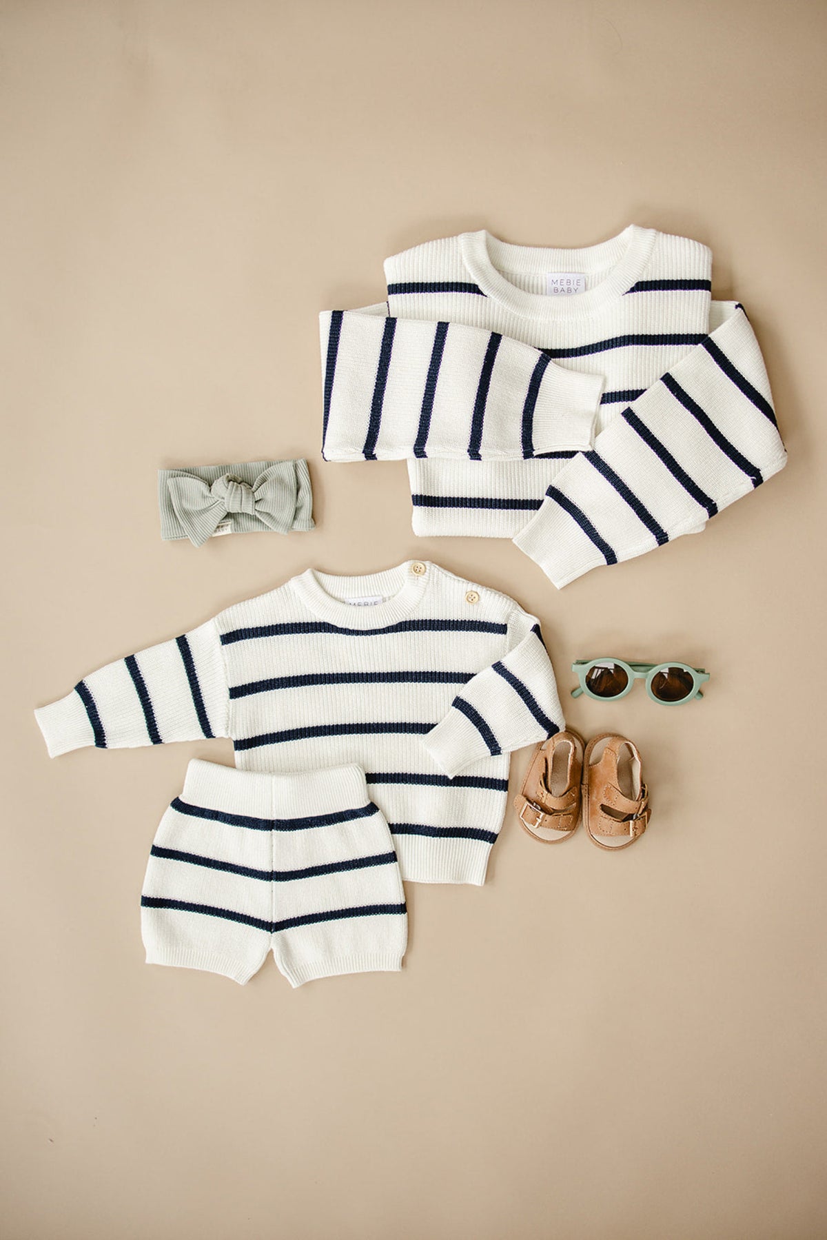 White and navy striped knit baby outfit with shorts, sweater, headband, sunglasses, and brown sandals on beige background.