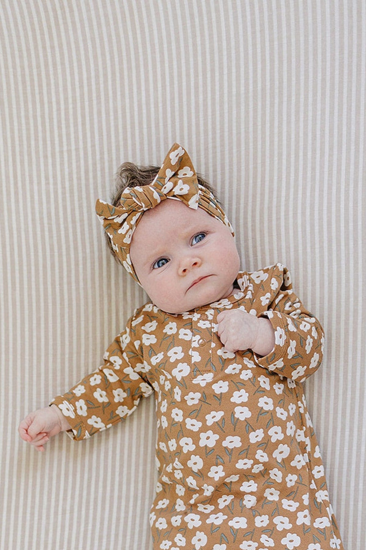 A baby with blue eyes lies on a striped background, wearing a floral mustard outfit and matching head wrap.