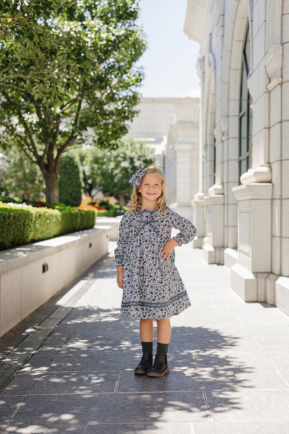 A young girl wearing a floral dress and matching headband stands smiling on a sunlit stone walkway.