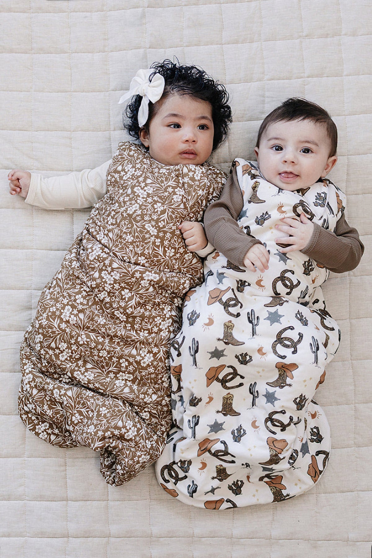 Two babies lie side by side on a textured, light-colored blanket, each wearing a different patterned sleep sack made from soft fabric.