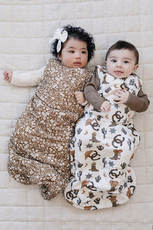 Two babies lie side by side on a textured, light-colored blanket, each wearing a different patterned sleep sack made from soft fabric.