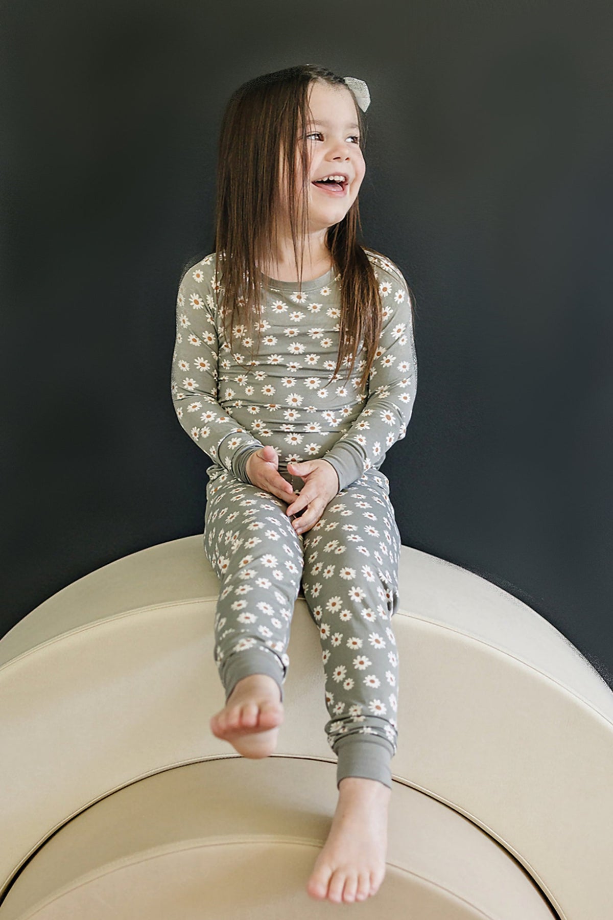 A child in a light green daisy-patterned pajama set sits smiling on a curved, beige surface.