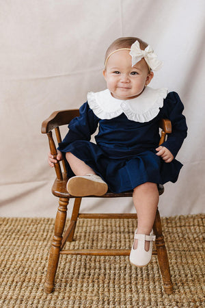 A cheerful baby sits on a wooden chair, wearing a navy dress with a white Peter Pan collar.