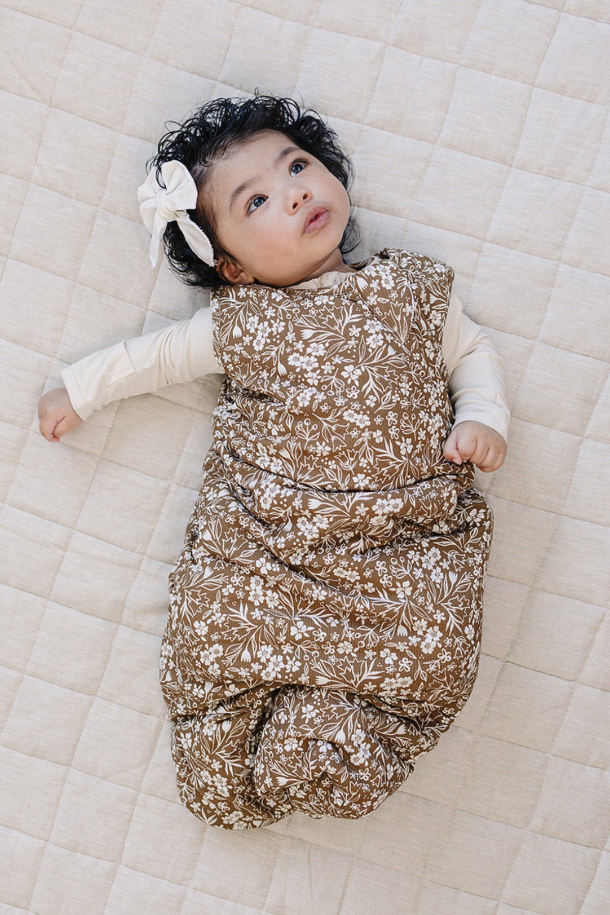A baby lies on a textured beige mat, wearing a brown floral sleep sack made from soft fabric. The infant has curly hair and a white bow, looking up thoughtfully.