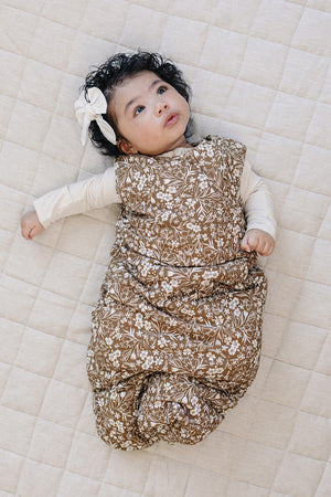 A baby lies on a textured beige mat, wearing a brown floral sleep sack made from soft fabric. The infant has curly hair and a white bow, looking up thoughtfully.