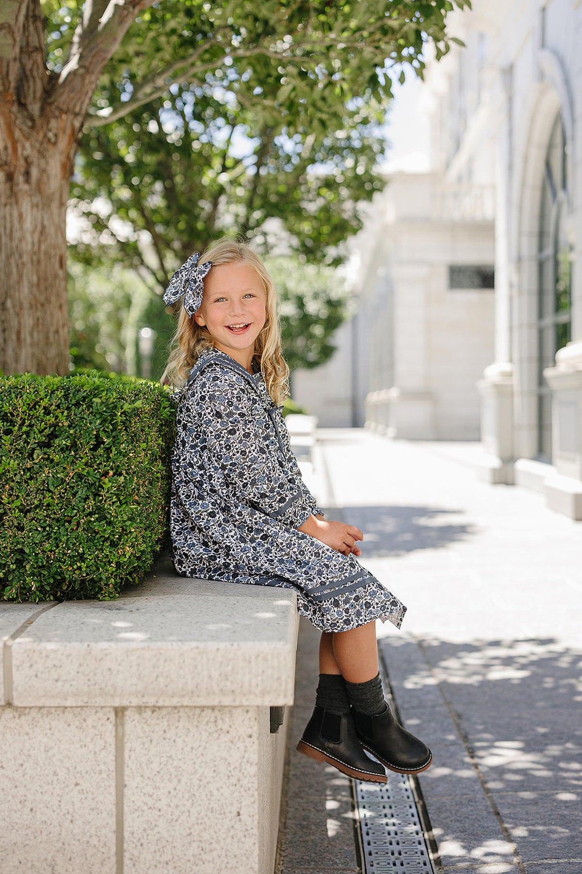 A young girl in a blue floral dress with a matching headband sits on a stone ledge outdoors.