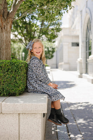 A young girl in a blue floral dress with a matching headband sits on a stone ledge outdoors.