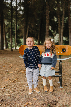 Two children stand together in a forest setting, holding hands. The boy wears a navy striped long sleeve shirt, while the girl is in a sweater and denim skirt, both.